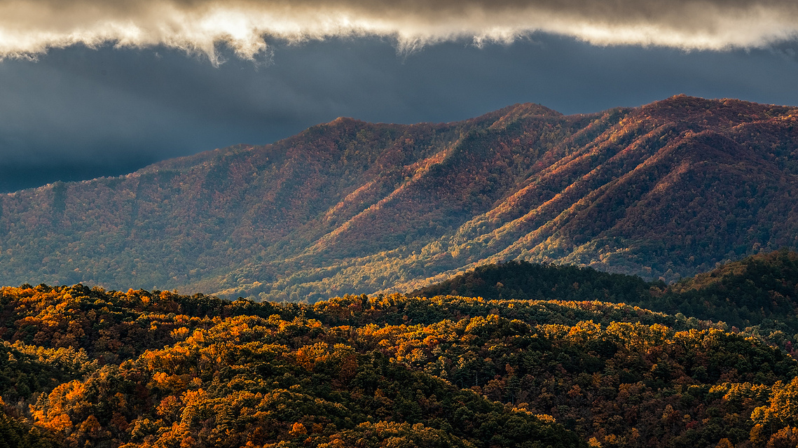 Golden light breaks through dark clouds to illuminate autumn-covered ridges in Great Smoky Mountains National Park, casting dramatic highlights across the layered forested slopes.