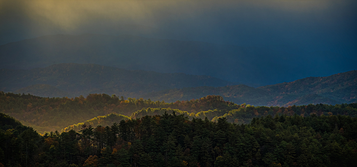 Golden morning light breaks through the misty fog to highlight forested ridges in Great Smoky Mountains National Park, casting a soft glow across the autumn landscape beneath deep blue shadows.