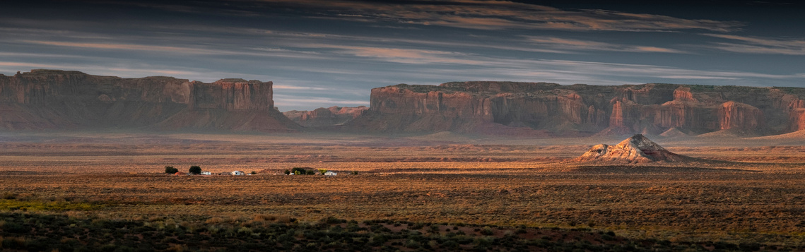 A small homestead rests quietly on the vast desert floor near Monument Valley, Utah, dwarfed by towering red rock mesas and bathed in the soft light of dusk beneath a streaked sky.