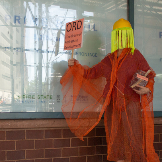 artist wearing construction hat with neon fringe and holding  ORD [The Oracle of Real-estate Development] sign