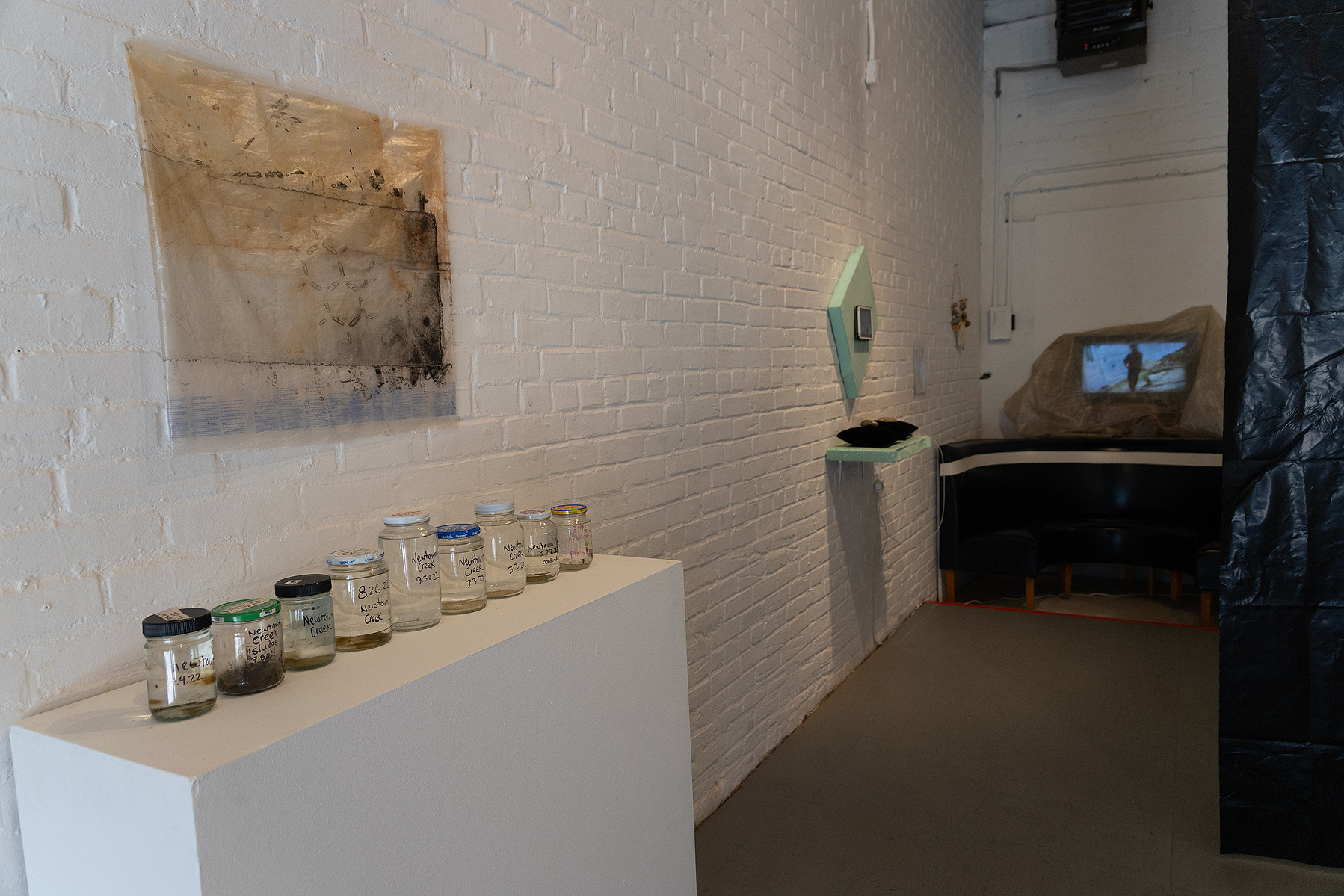 Works from Priscilla Stadler's SLUDGE project, an art installation created in response to research about the toxic waterway Newtown Creek, NYC. On the left, "Troubled Waters 11" on wall, "Samples" is jars of Creek water on pedestal.