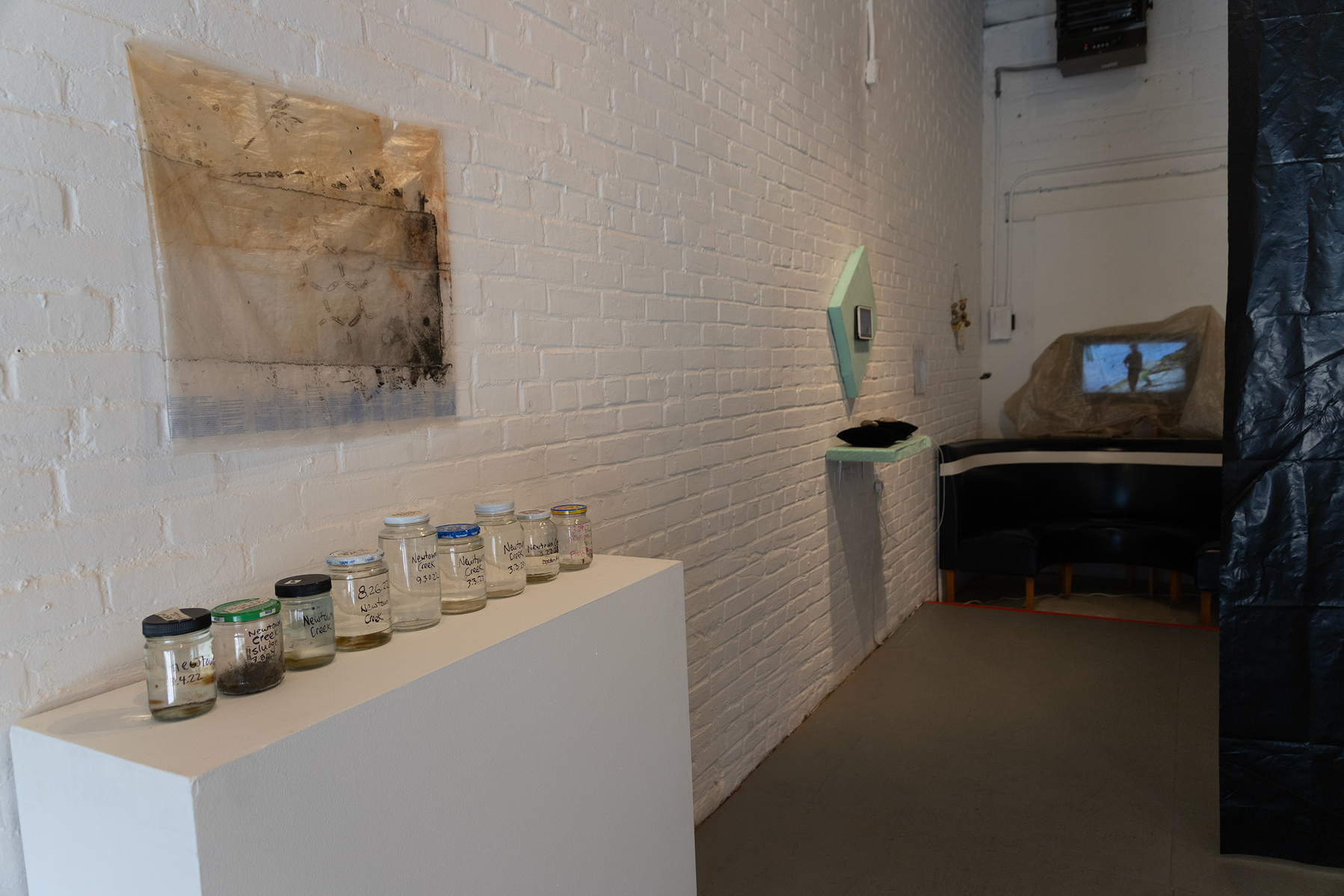 Works from Priscilla Stadler's SLUDGE project, an art installation created in response to research about the toxic waterway Newtown Creek, NYC. On the left, "Troubled Waters 11" on wall, "Samples" is jars of Creek water on pedestal.