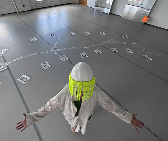 Artist Priscilla Stadler dressed as the Oracle  wearing a lab coat and a white construction helmet with fringe covering her face. With outstreteched arms she stands in front of a 15-foot transparent plastic cut-out she made of Newtown Creek