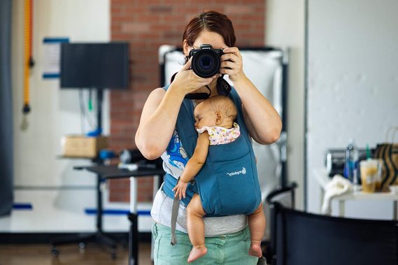Nekoda Mattox holding a canon eon r5 to her face in a photography studio while wearing a blue ergo embrace baby carrier with a six month old baby girl.