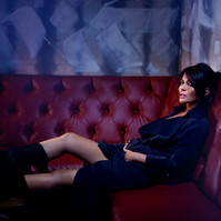 Editorial photo of actress Marisol Nichols posing on a red tufted couch with legs up on a chair, with tall black heeled boots and deep blue wrap dress. 