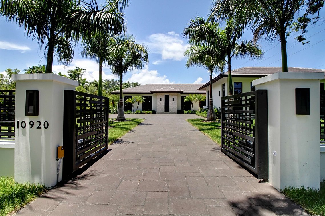 A striking modern estate with a gated entrance, lush tropical landscaping, and a sleek driveway leading to the main residence.