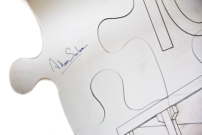 A detail of a wall hung sculpture made of aluminum forms 4 pieces of an oversized jigsaw puzzle with a painted drawing of a building construction on it.