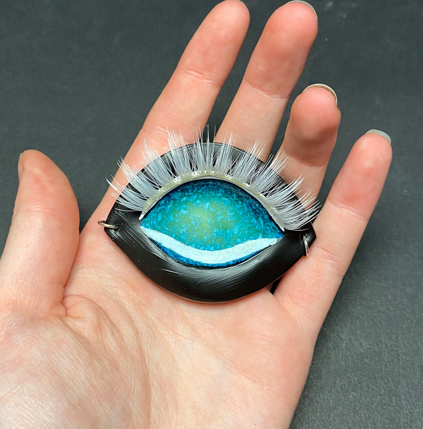 A ceramic pendent of an eye. The eye base is black with a bright blue iridescent core. Adorned with white eyelashes and liner.