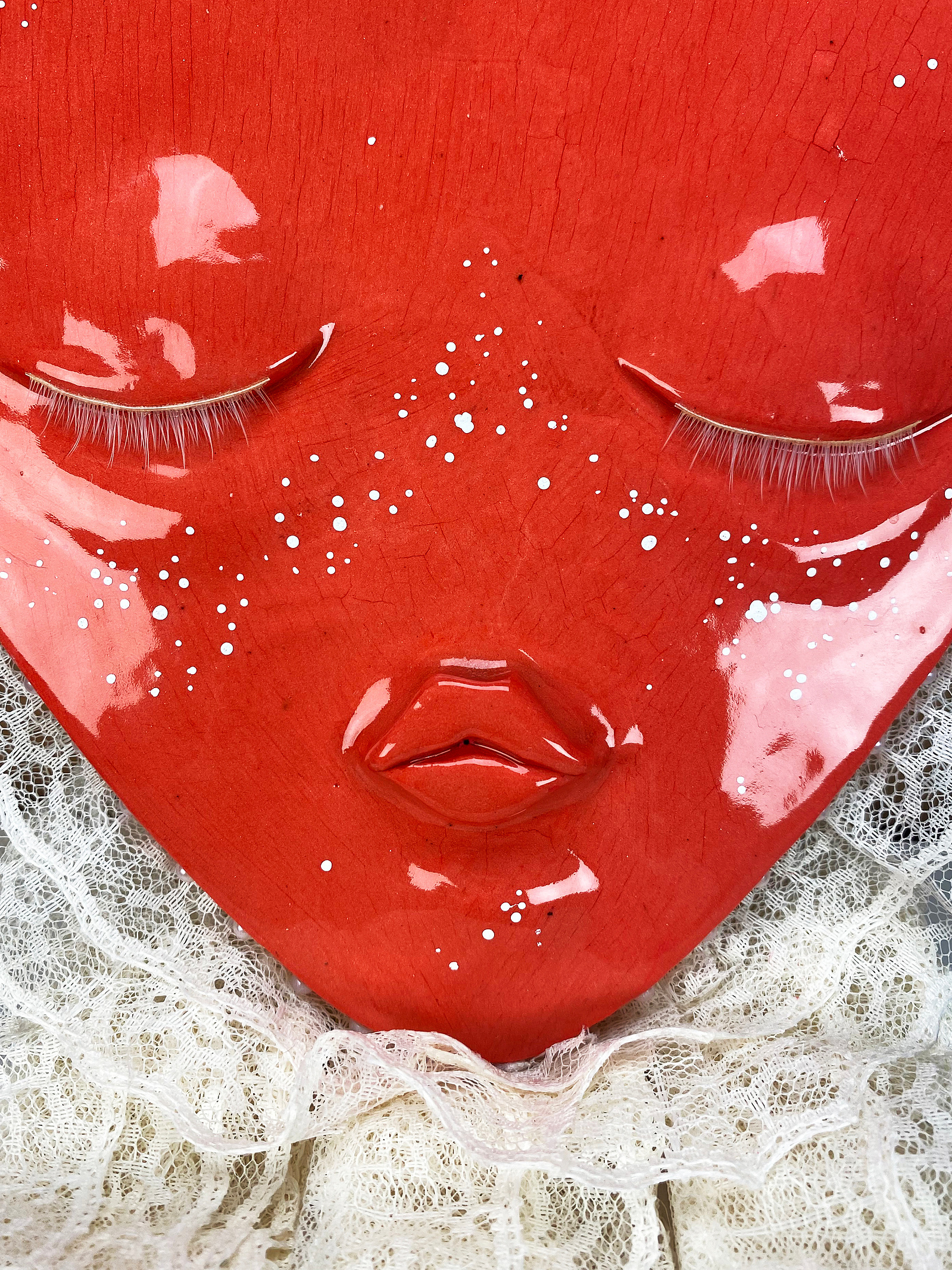 A ceramic heart shaped face with closed eyes and a pouted mouth. Adorned with white eyelashes, white freckles, a white lace trim and hung with a ribbon. Pearls line the sides.