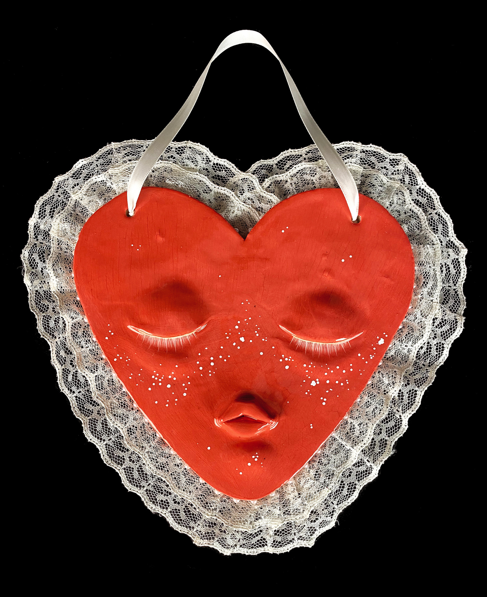 A ceramic heart shaped face with closed eyes and a pouted mouth. Adorned with white eyelashes, white freckles, a white lace trim and hung with a ribbon. Pearls line the sides.