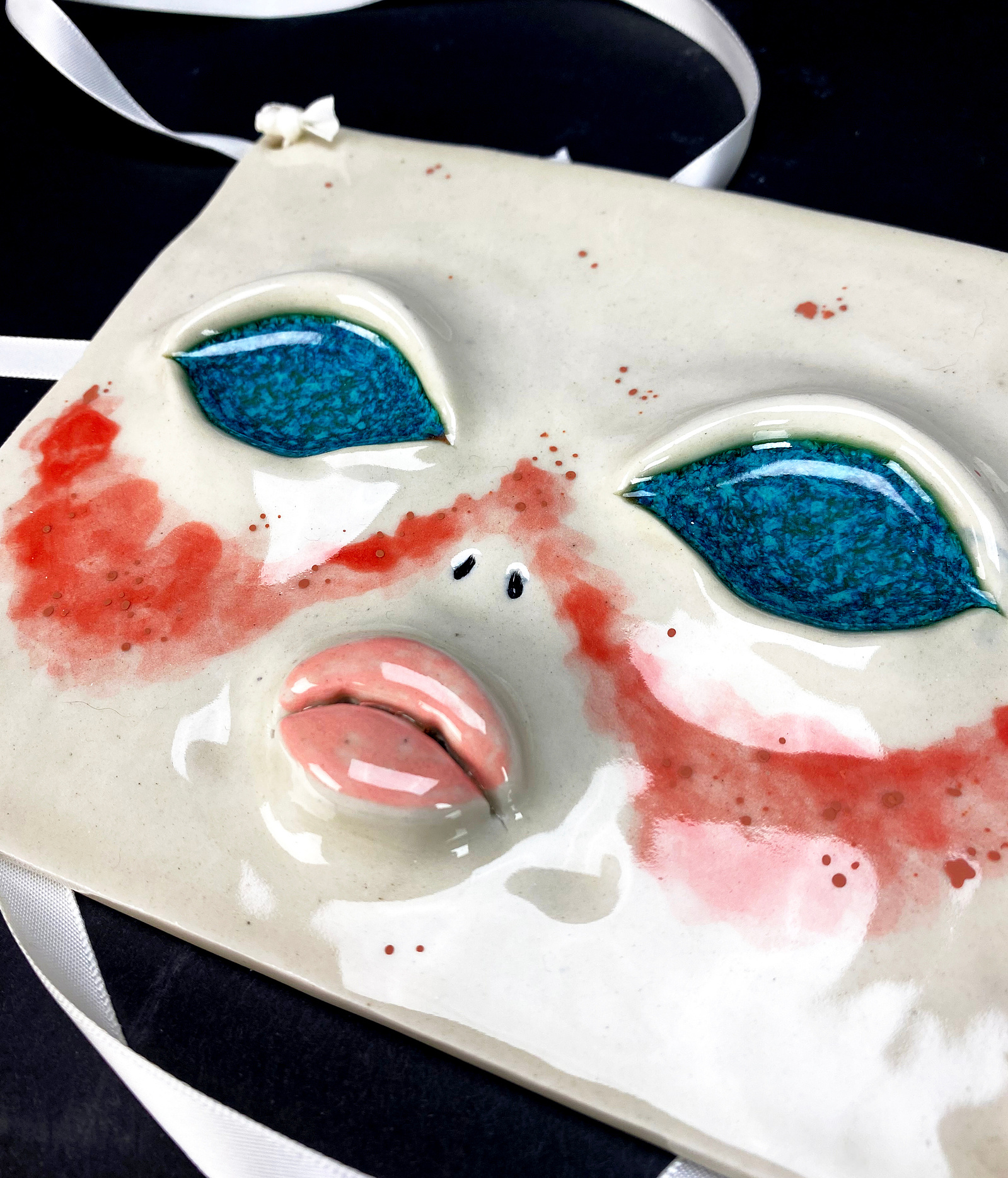 A ceramic face mask using ribbon as the strings. Bright blue iridescent eyes with pouty lips and blush.