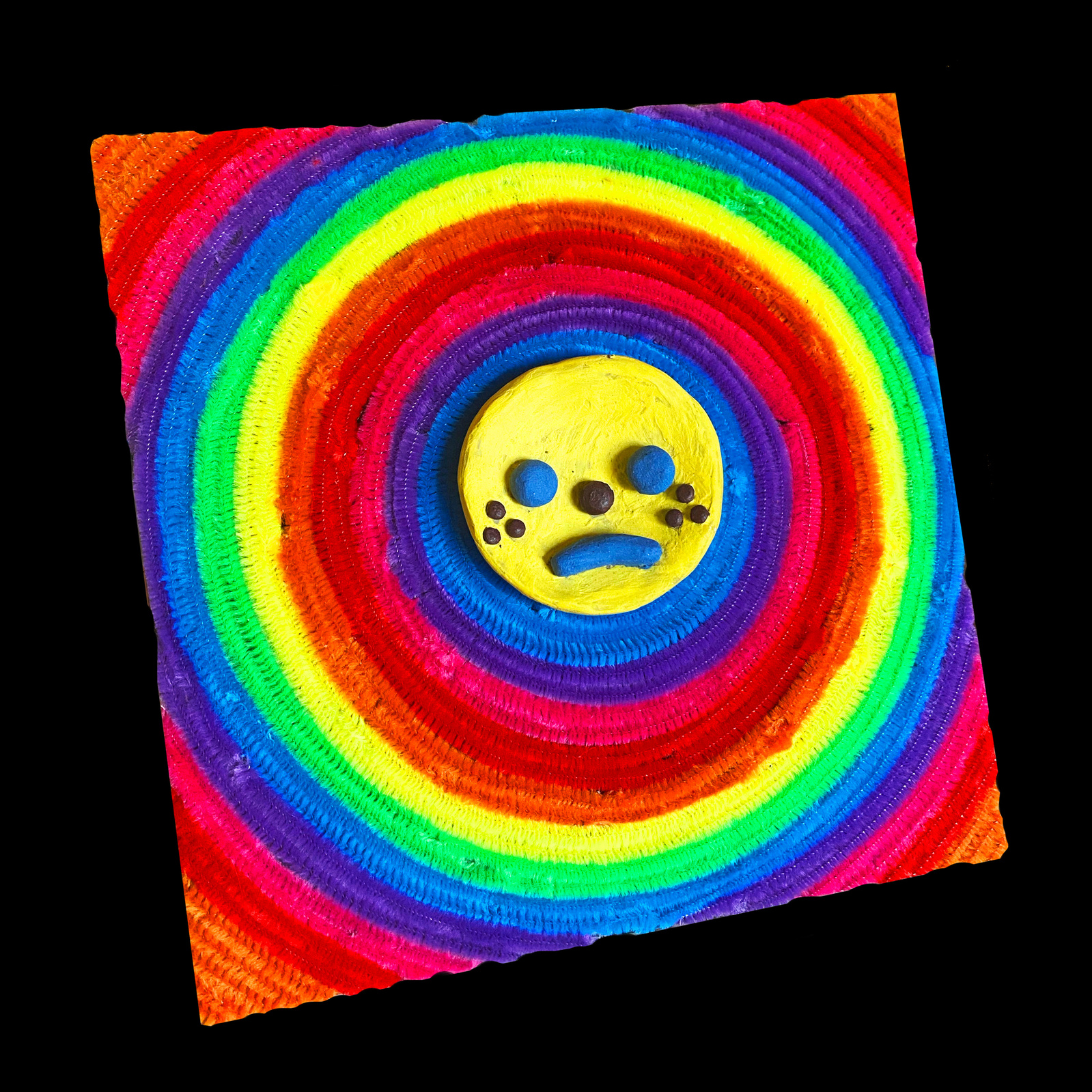 A multi media piece. A ceramic frowny face surrounded by color coded pipe cleaners on canvas.