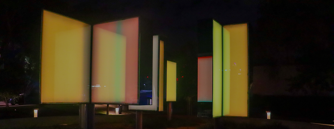 A picture of a light sculpture in West Hollywood. The sculpture is made up of rectangular panels placed in opposite angles to each other, with colors of yellow, pink, white, and green.