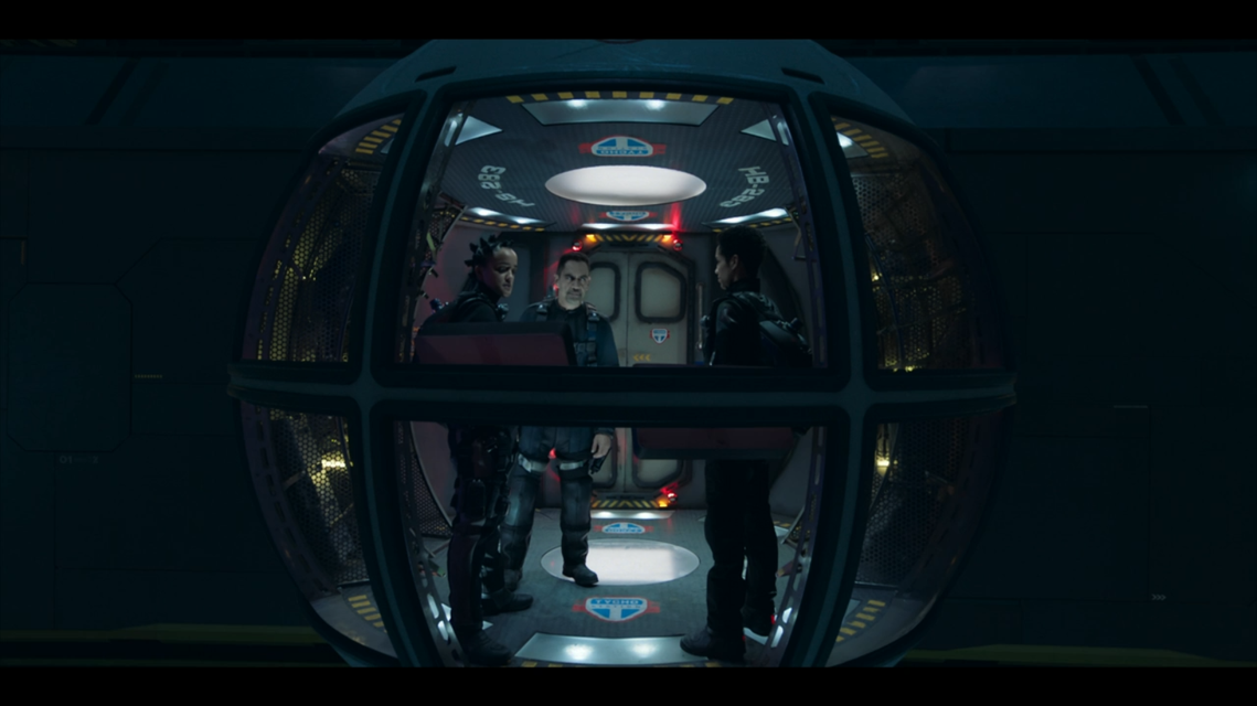3 actors stand in a spherical pod space shuttle. There are 8 curved strut that devides the pod into sections. The front 3 are floor to ceiling windows. The rear 3 are a door in the centre with walls. There is a control console and industrial style storage