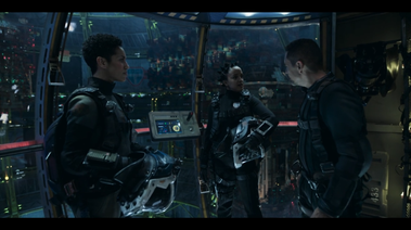3 actors stand in the pod, 2 of which are in spacesuits.  Behind can be found handrails, some of the console details, and a space station in the background