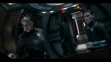 3 actors stand in the pod. behind them you can see the under-lit walkway of the dock, and the door leading to the rest of the space station.