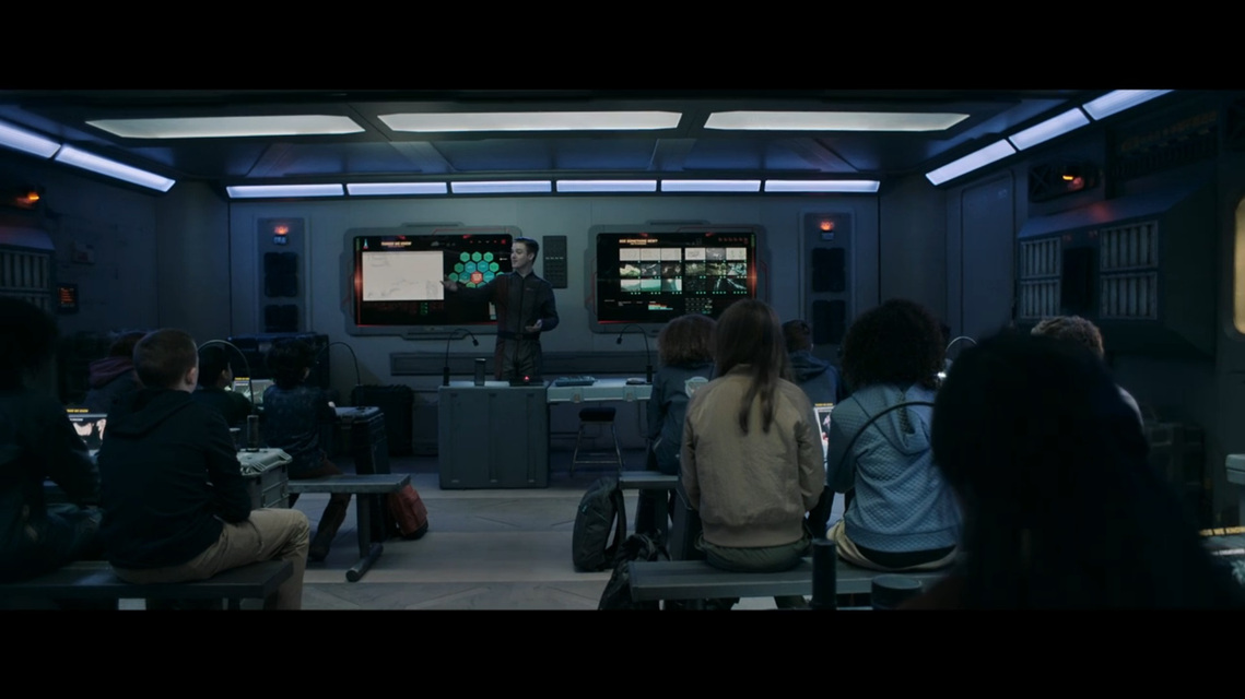 A teacher stands at the front of a futuristic classroom in front of many students (all actors). There are 2 large screens, on the wall behind the teacher, and the students sit at makeshift desks made of creates and containers in front of benches.