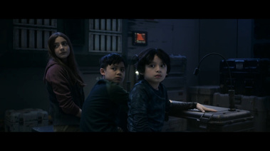 3 children sit at a makeshift desk. There is a console on the wall behind them, with red lights. There are giant vents on the walls.