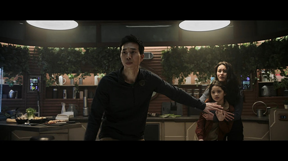 A man stands in front of and guarding his wife and daughter. Behind them is a kitchen, built partially across 3 walls of a hexagonal room. There is a cantilevered dining table with stools, a sink, a cooking space and a fridge, all with storage below. 