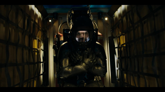 Man in a spacesuit is strapped into a fighter-jet style chair in a very compact space. The walls look like a corrugated shipping container and there are pipes and straps on the walls.