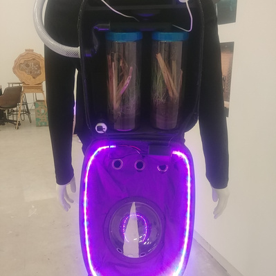 The backpack device on a mannequin. The backpack is fully open, showing the contained ecosystems, pipes, hoses, batteries, fans, wires and LEDs.