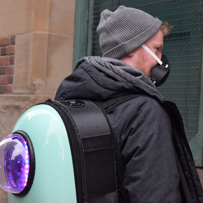Man stands in an alley looking over his shoulder. He wears a backpack device which has glowing lights seen through a window.