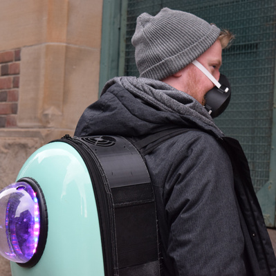 Man stands in an alley looking over his shoulder. He wears a backpack device which has glowing lights seen through a window.