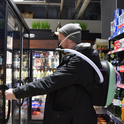 Man in a corner shop is opening a large fridge. He wears a backpack device which has a hose connected to a facemask on his face.