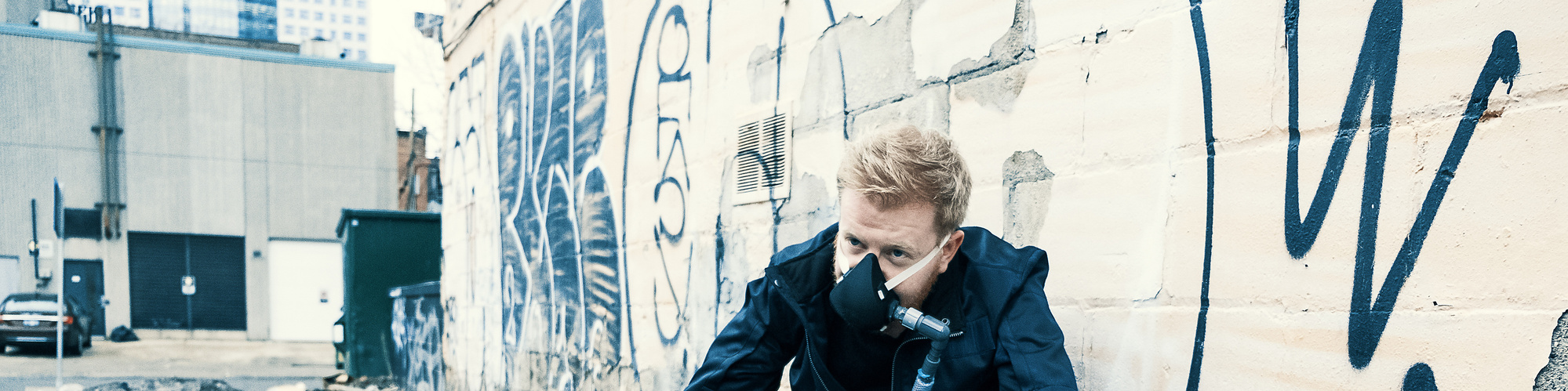 Man squatting by a graffiti covered wall. He wears a mask, which is connected by a hose to a backpack which has glowing lights seen through a window.