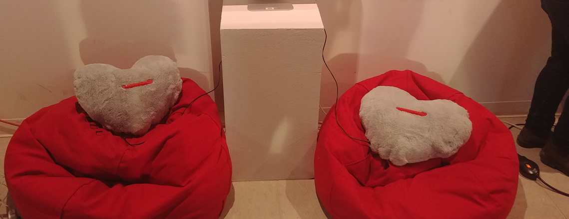 2 grey pillows each sit on 2 red bean bags on opposite sides of a plinth. Both pillows have red plastic strips in the middle.