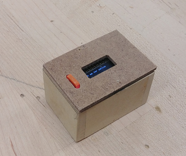 Small handmade wooden box, with a small orange button and LED screen with writing on it .