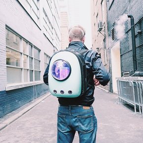 Man stands in an alley facing away. He wears a backpack device which has glowing lights seen through a window. There are containers which can bee seen to contain plant matter.