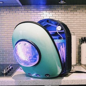 The backpack device sitting on a kitchen counter partially open. Inside there are wires, fans, batteries and LED lights, and 2 containers containing plant based ecosystems.