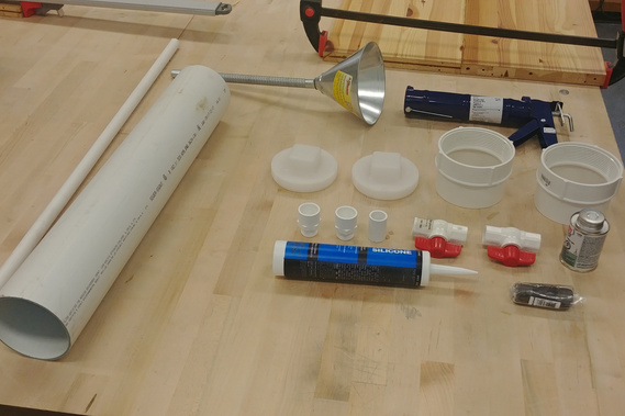Components made to build the prototype on a wooden workshop table. Included: 2 lengths of different width PVC piping, a metal funnel, plastic caps, threaded pipe ends, ball stop valves, thread-maker, pipe ends, adhesives and silicone. 