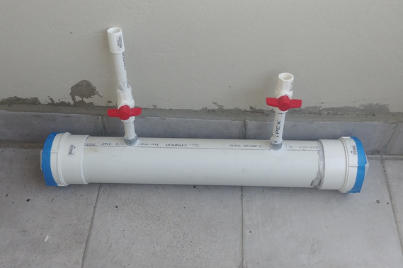 Prototype for a testing environment. It is made of piping and plumbing fixtures. A wide PVC pipe is capped at both ends, and has 2 smaller pipes entering from the top, both with ball stop valves.