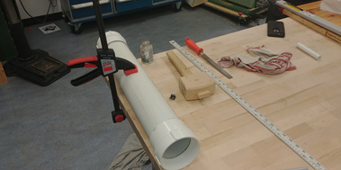 Work-in-progress of the construction. PVC pip with fitted thread caps, is clamped to a workshop table. There are tools on the table, including a wooden mallet, a ruler and a file.