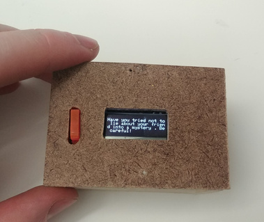 Top-down view of the small wooden box with the orange button. The screen on the top reads 