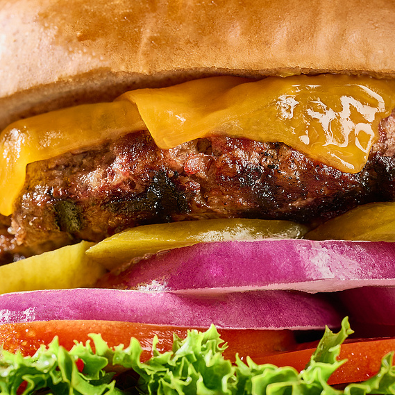 Macro shot of burger. Burger fills the entire frame and layers are visible- bun, melted cheese, patty, pickles, red onion, tomato, and green leaf lettuce, bottom bun. Image by Megan Morello San Diego Food and Lifestyle Photographer.