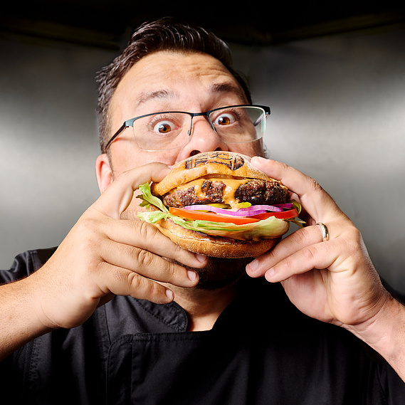 Chefs portrait. Man wearing glasses and black chef's coat, biting into large burger with wide eyes. Image by Megan Morello San Diego Food and Lifestyle Photographer.