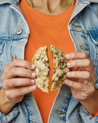  Woman wearing orange shirt and denim jacket. Her hands are splitting a cookie in half so the chocolate chips inside can be seen. Image shot by Megan Morello, San Diego and Southern California Food and Photographer.