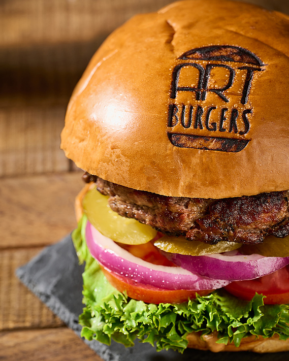 3/4 angle shot of burger. Restaurant logo branded into top bun. Colorful layers of brown, yellow, purple, red visible. Burger is on slate board on top of wood surface. Image by Megan Morello, San Diego food and lifestyle photographer.
