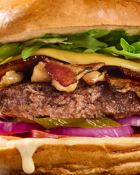 Macro shot of burger filling the frame. Layers Visible-bun, arugula, gouda cheese, maple bacon, roasted garlic cloves, patty, pickles, red onion, tomato, sauce, bun. Image by Megan Morello San Diego Food and Lifestyle Photographer.