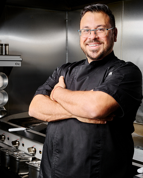 Chef standing in his sparkling clean stainless steel industrial kitchen. Chef is wearing black chef's coat and glasses, smiling into the camera with arms folded across chest. By Megan Morello, San Diego food and lifestyle photographer.