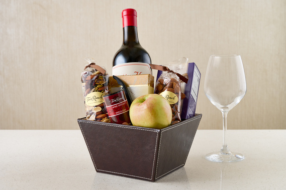 Corporate looking gift box filled with bottle of red wine, various packages of nuts, and an apple. Wine glass also in frame. Beige background. By Megan Morello Commercial food photographer and lifestyle photographer in San Diego, Southern California.