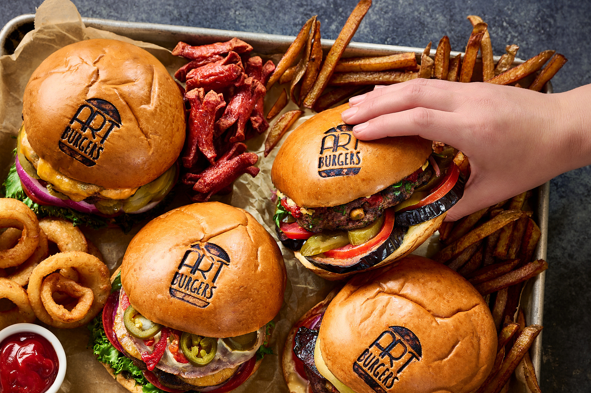 Family style tray of burgers, fries, onion rings, and sausages. Hand reaching in and grabbing a burger. Image by Megan Morello San Diego Food and Lifestyle Photographer.