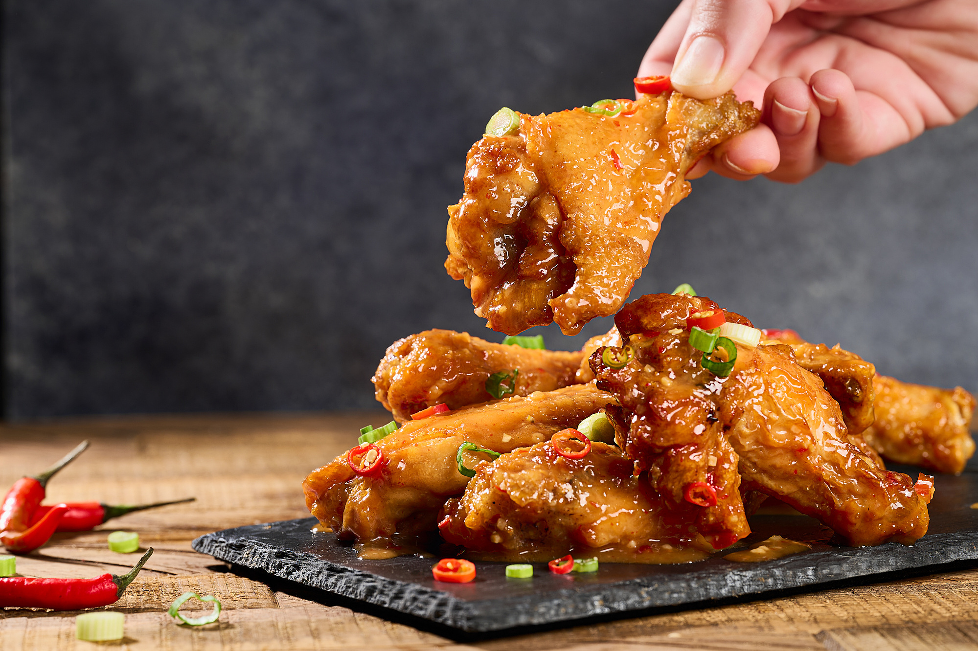 Wings on a slate board with a spicy sauce and garnished with green onion and red chili rounds. Hand reaching for wing. Image by Megan Morello San Diego Food and Lifestyle Photographer.