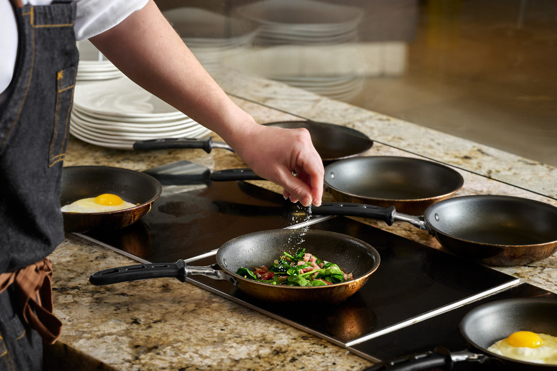 Close up of chef's hand sprinkling salt into a pan of sautéing vegetables. Fried eggs visible in other 2 pans in frame. Image by Megan Morello Commercial food photographer and lifestyle photographer in San Diego, Southern California.