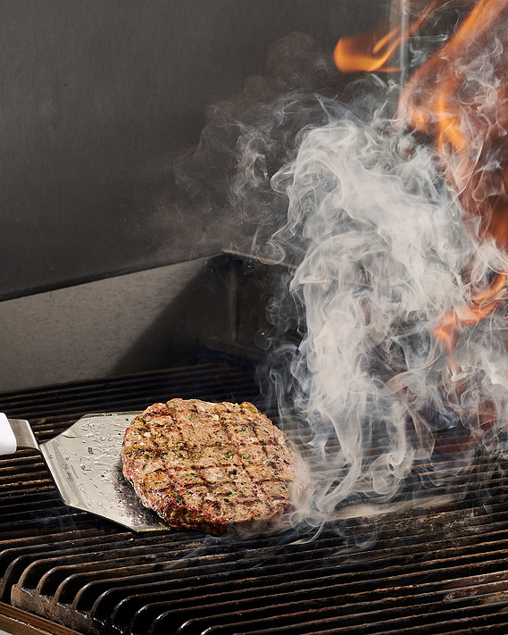 Burger parry cooking on the grill. Beautiful has marks visible on the burger. Smoke and fire rise from the grill. Image by Megan Morello San Diego Food and Lifestyle Photographer.