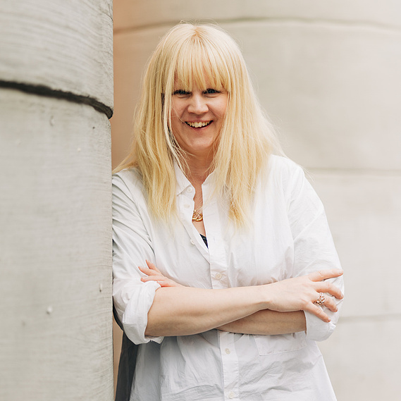 Åsa Törnquist leaning against a cement wall. The  image has a soft light. She is blond with her signature hairstyle - a fringe. She has a white shirt on. 