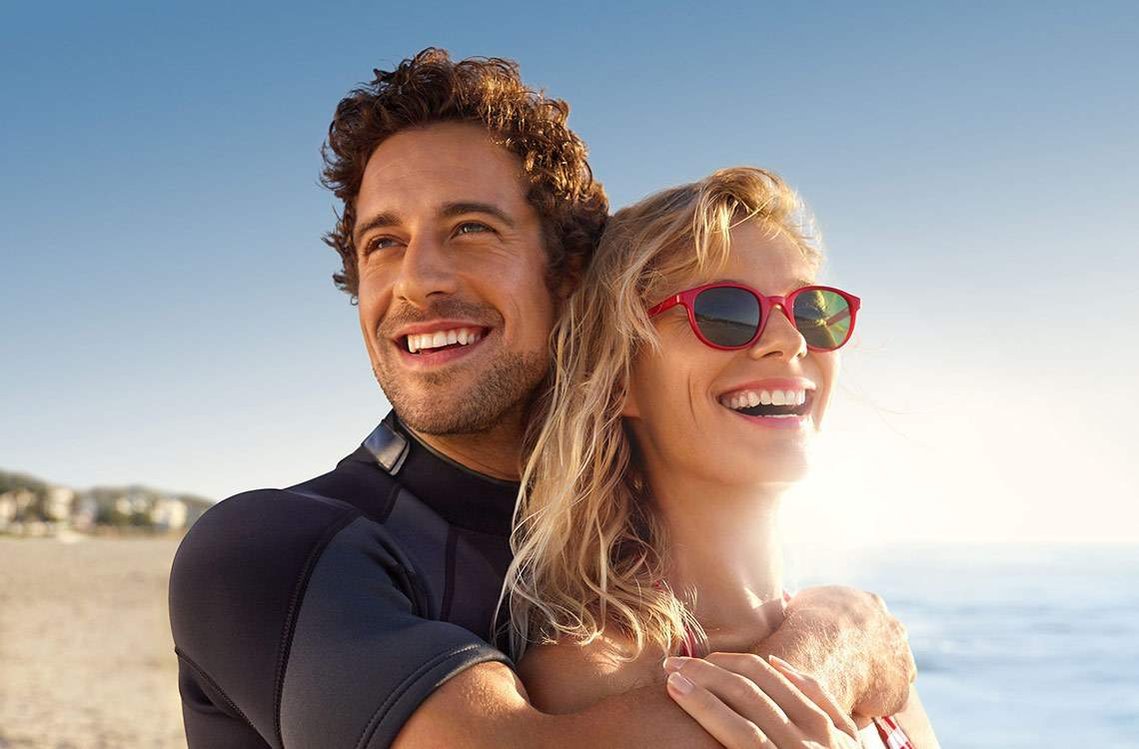 Ein Mann in Neoprenanzug hält eine Frau mit roter Sonnenbrille im Arm